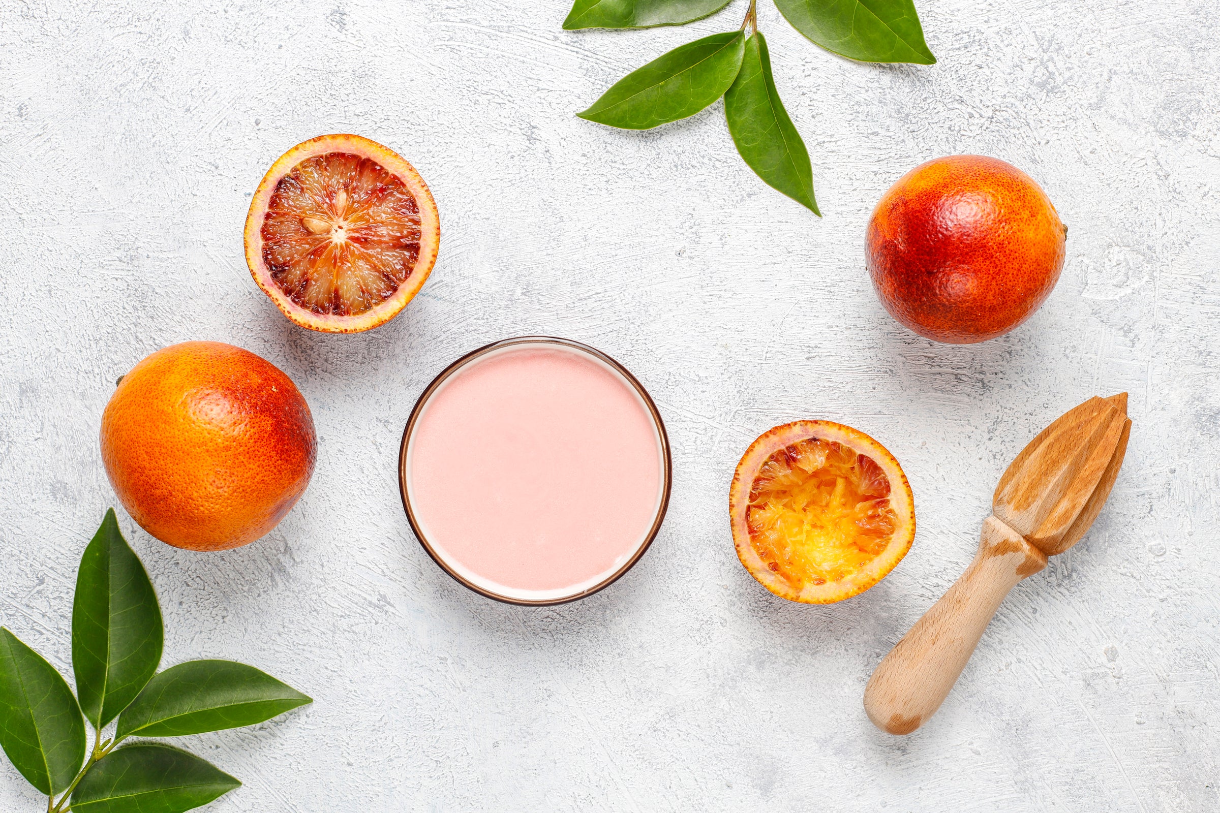 Oranges sanguines entières et coupées, un bol de jus rose, et un presse-agrumes en bois sur fond gris.